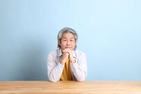 The senior Asian man with grey shirt sitting at the working desk with the blue background.
