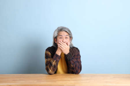 The senior Asian man wearing the yellow plaid shirt sitting at the working desk with blue background.の写真素材