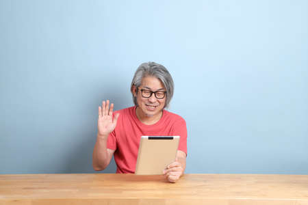 The senior Asian man with colorful shirt sitting at the desk with blue background.の写真素材