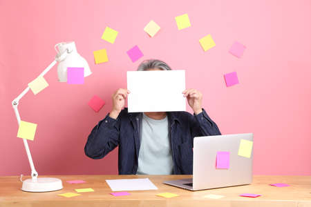 The senior Asian man sitting at working desk with a lot of paper work against the pink background.