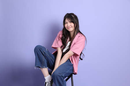 The cute young Asian girl with casual clothes sitting on the purple background.