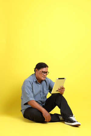 The adult Asian man with blue shirt sitting on the yellow background.