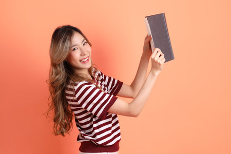 The young Asian woman in casual clothes with gesture of holding a book on the orange backgroundの写真素材