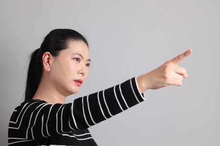 The 40s East Asian woman in the casual clothes with gesture of pointing on the grey background.