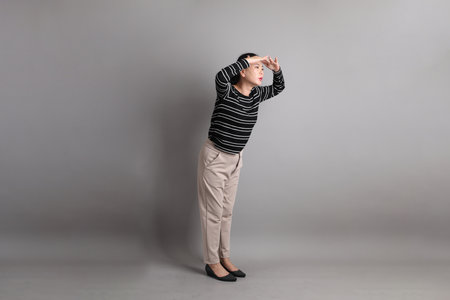The 40s East Asian woman in the casual clothes with gesture of peering into distance, looking far away on the grey background.の写真素材