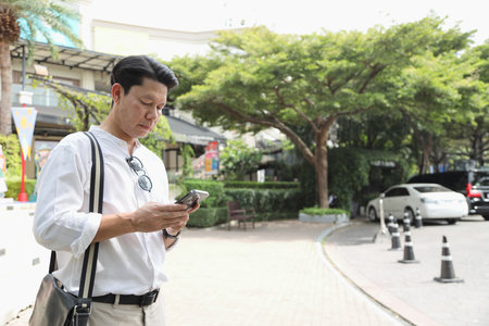 The 40-year-old Asian businessman walking in the parking lot while using a smartphone during the daytime.の写真素材