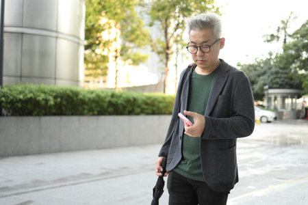 A senior Asian man using a smartphone is walking in the business area.の写真素材