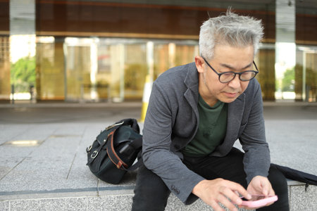 A senior Asian businessman using a smartphone is sitting near the outdoor stairs.の写真素材