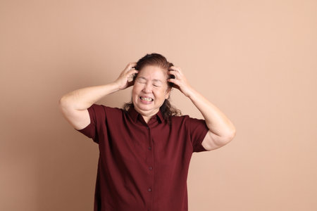 Senior East Asian woman with a deep red shirt standing against a brown background.の写真素材