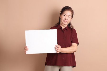 Senior East Asian woman with a deep red shirt standing against a brown background.の写真素材