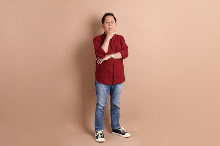 Senior East Asian man with a deep red shirt standing against a brown background.の写真素材