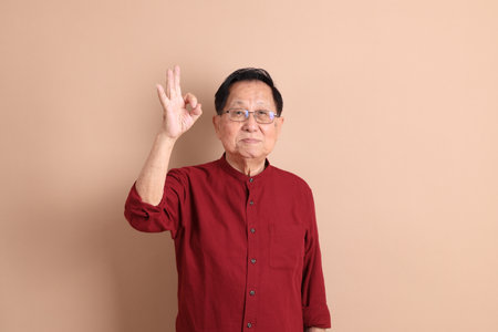 Senior East Asian man with a deep red shirt standing against a brown background.の写真素材