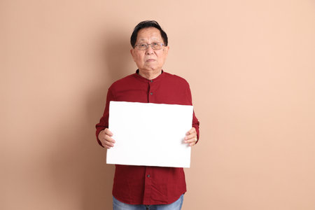 Senior East Asian man with a deep red shirt standing against a brown background.の写真素材