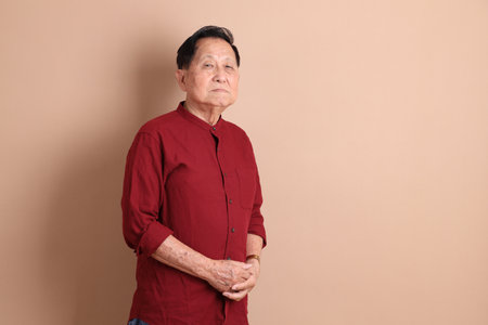 Senior East Asian man with a deep red shirt standing against a brown background.の写真素材