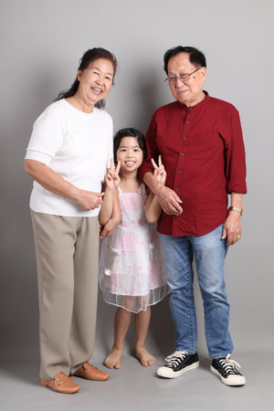 A senior East Asian grandparent with their granddaughter in casual clothes, standing against a grey...