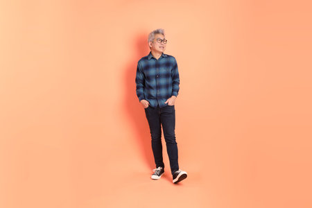 A senior East Asian man in a deep blue plaid shirt stands against a peach-pink background.の写真素材