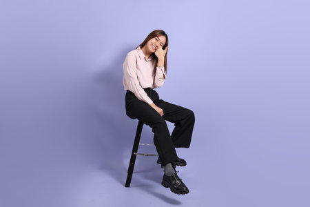 A young Southeast Asian woman in business clothes, sitting on a stool against a purple background.