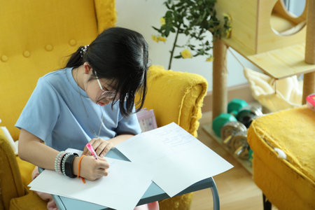 The East Asian girl is doing homework at home on the armchair.の写真素材
