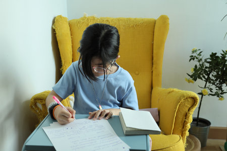 The East Asian girl is doing homework at home on the armchair.