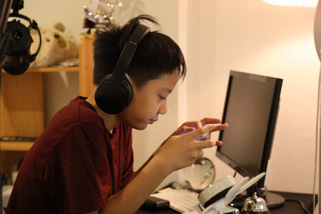 The boy is playing an online game on his smartphone at the computer desk.