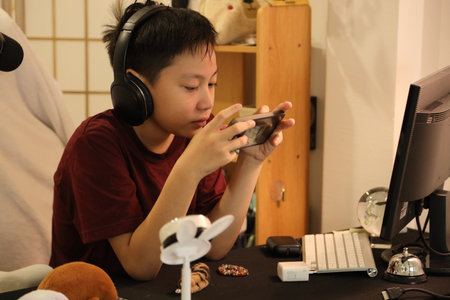 The Southeast Asian boy is playing an online game on his smartphone at the computer desk.