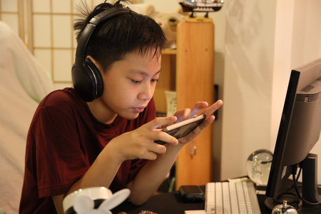The Southeast Asian boy is playing an online game on his smartphone at the computer desk.