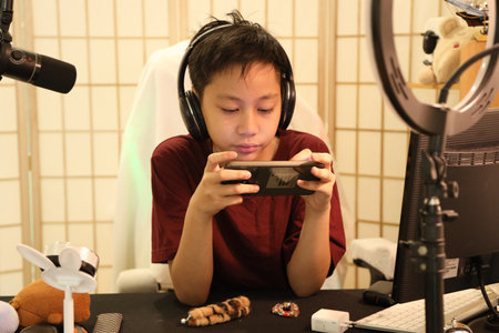 The Southeast Asian boy is playing an online game on his smartphone at the computer desk.
