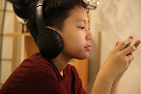 The Southeast Asian boy is playing an online game on his smartphone at the computer desk.
