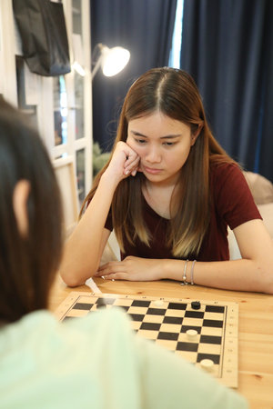 Two Southeast Asian teenage girls are playing a game of draughts together in the living room.