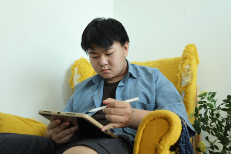 The Southeast Asian teenage boy is drawing a picture on a digital tablet in the armchair.の写真素材