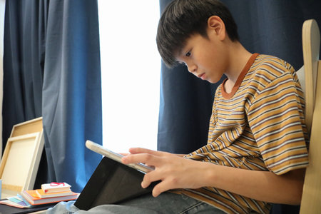The Southeast Asian boy is using a digital tablet in the living room.