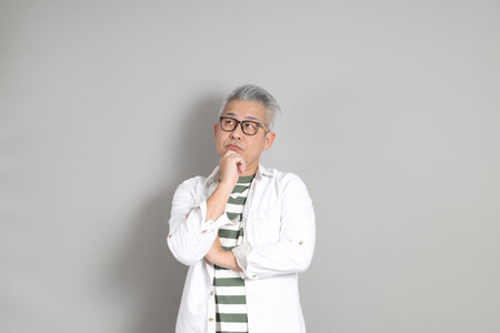 The portrait of an East Asian man with gray hair and glasses, dressed in smart casual clothes, standing against a gray background.の写真素材