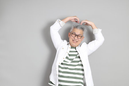 The portrait of an East Asian man with gray hair and glasses, dressed in smart casual clothes, standing against a gray background.の写真素材