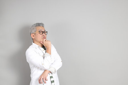The portrait of an East Asian man with gray hair and glasses, dressed in smart casual clothes, standing against a gray background.の写真素材