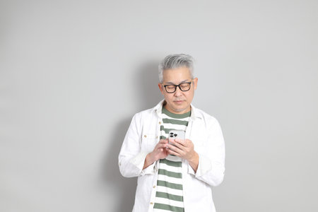 The portrait of an East Asian man with gray hair and glasses, dressed in smart casual clothes, standing against a gray background.の写真素材