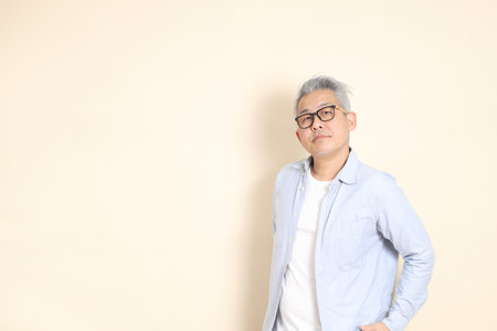 The portrait of an East Asian man with gray hair and glasses, dressed in smart casual clothes, standing against a brown background.の写真素材