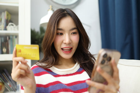 The East Asian woman is using a smartphone to do financial tasks or shop online with a credit card at home in the living room.の写真素材