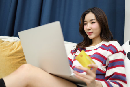 The beautiful East Asian woman is using a credit card to do financial tasks on her laptop in the living room.の写真素材