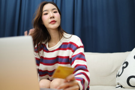 The beautiful East Asian woman is using a credit card to do financial tasks on her laptop in the living room.の写真素材