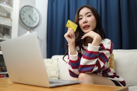 The beautiful East Asian woman is using a credit card to do financial tasks on her laptop in the living room.の写真素材