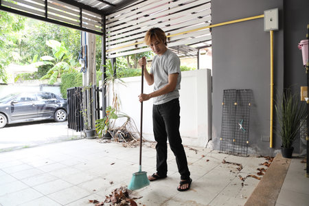 The man is cleaning and sweeping the leaves in front of the house.