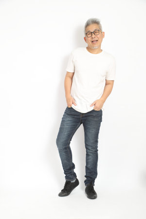 Friendly mature East Asian man in a white t-shirt and blue jeans standing with hands in pockets and...