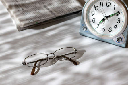 Eyeglasses, table clock and newspaper on a desk by the windowの写真素材