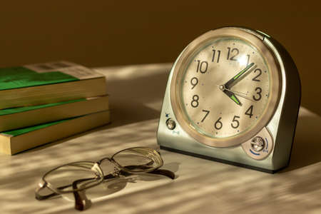 Eyeglasses, table clock and books on the desk by the window in the eveningの写真素材
