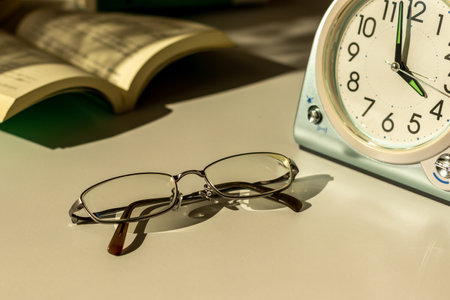 Eyeglasses, table clock and books on the desk by the window in the evening