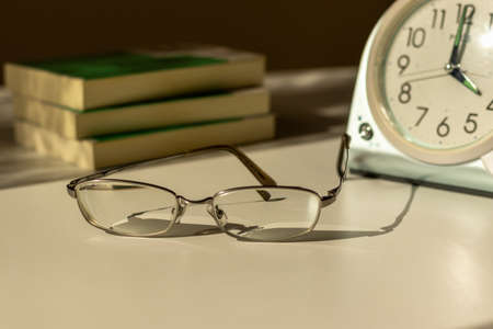 Eyeglasses, table clock and books on the desk by the window in the eveningの写真素材