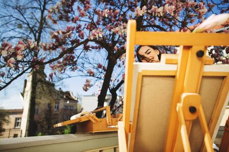 Young brunette woman artist paints a picture near the magnolia tree and various art equipment. An easel in the foreground.の写真素材