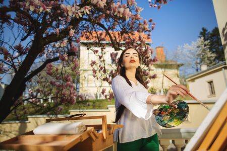 Young brunette woman artist holding in hands a brush and palette. Girl standing near the magnolia tree and various art equipment.の写真素材