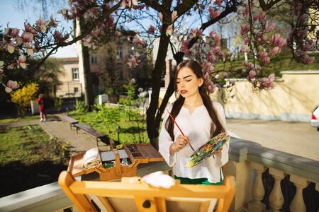 Young brunette woman artist holding in hands a brush and palette. Girl standing near the magnolia tree and various art equipment.の写真素材