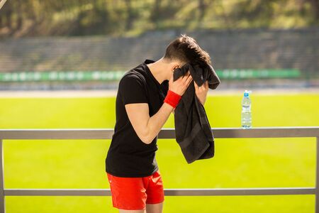Young sporty man resting and wiping his sweat with a towel after workout sport exercises outdoors on the background of green grass.の写真素材
